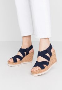 Navy wedged sandals with cork heel, elastic crisscross straps, open toe design, and rubber sole. Curved wedge adds height.