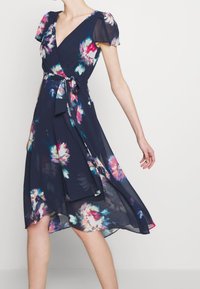 Navy floral wrap dress dengan lengan puff pendek, sabuk ikat, dan rok melayang yang menampilkan pola bunga cerah dalam nuansa pink dan teal.