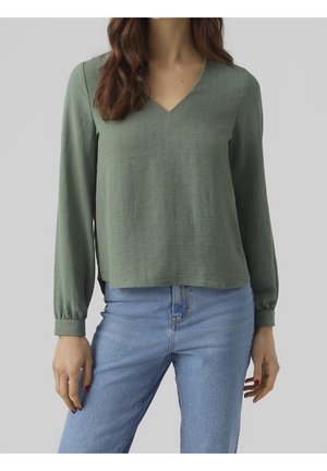 Kvinde iført en olivengrøn bluse med lange ærmer og V-hals, kombineret med lyseblå højtaljede jeans, foran en grå baggrund.