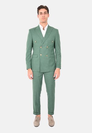 Suit verde doppiopetto con bottoni dorati, tessuto strutturato, pantaloni slim-fit e risvolti, abbinato a scarpe di colore chiaro.