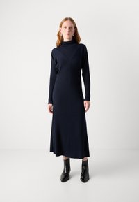 Vestido de punto acanalado azul marino con cuello alto simulado, mangas largas y una silueta de longitud media, combinado con botas negras hasta el tobillo sobre un fondo liso.