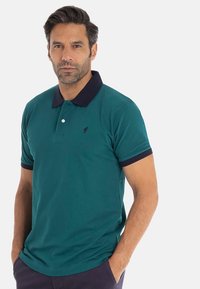Homme portant un polo teal avec un col et des bords de manches sombres, se tenant les mains dans les poches devant un fond uni.