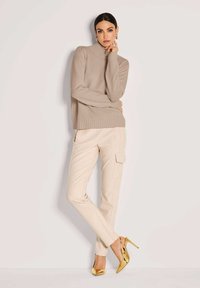 Frau in einem beigen Rollkragenpullover, cremefarbenen Cargo-Hosen und goldenen High Heels, die mit einer Hand ihr Gesicht berührt.