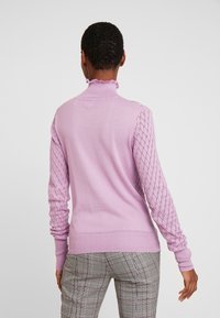 Personne aux cheveux courts portant un pull lavande aux manches texturées et un pantalon à carreaux gris à motifs, vue de dos sur un fond blanc.