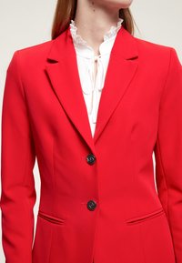 Röd kavaj med skräddarsydd passform, enkel knappstängning, två framfickor och eleganta slag. Under finns en vit blus med rynkad halsringning.