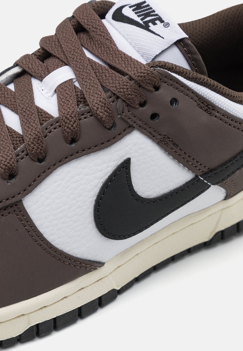 Basket en cuir marron avec panneau en textile blanc, logo Swoosh noir, lacets marron et semelle en caoutchouc crème. Détails de surface texturée visibles.