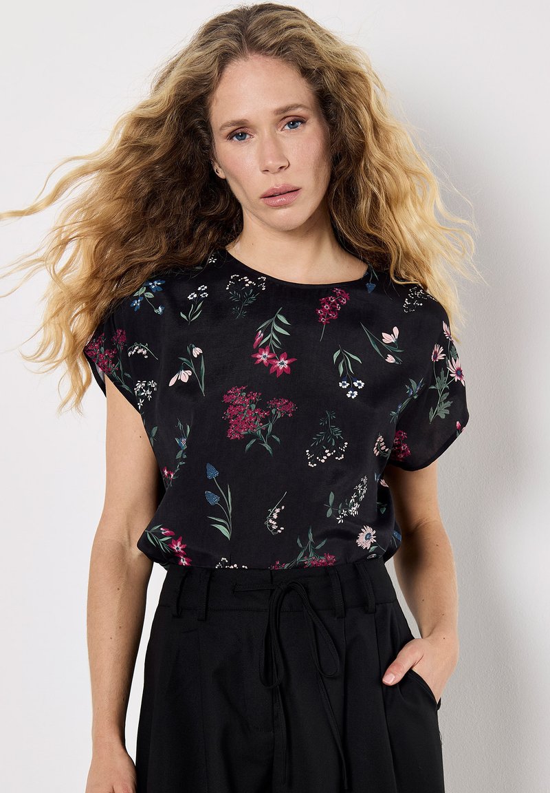 Haut noir à manches courtes avec imprimé floral en rouge, bleu et blanc. Tissu doux, encolure ronde, coupe ample, porté avec un pantalon noir taille haute.