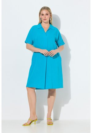 Kvinde med blondt hår iført en knælængde, lys blå kjole og gule hælede sandaler, stående mod en hvid baggrund.