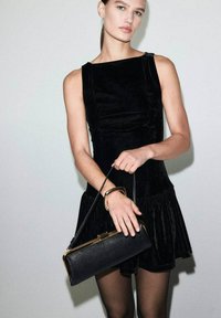 Kobieta w bezrękawnikowej sukience z czarnego aksamitu i przezroczystych czarnych rajstopach, trzymająca czarną torebkę z złotymi akcentami na jednolitym jasnym tle.