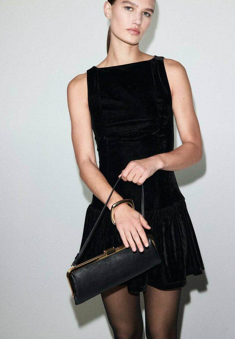 Kobieta w bezrękawnikowej sukience z czarnego aksamitu i przezroczystych czarnych rajstopach, trzymająca czarną torebkę z złotymi akcentami na jednolitym jasnym tle.
