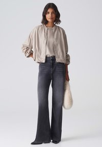 Hellbeige Bomberjacke mit strukturiertem Finish, kombiniert mit hoch taillierten, ausgestellten dunkelgrauen Jeans. Abgerundet mit einer Clutch aus Kunstfell.