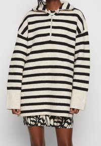 Personne portant un pull en maille beige et noire à larges rayures horizontales avec une fermeture éclair sur la moitié, et une jupe ou une robe noire et blanche à motifs en dessous.