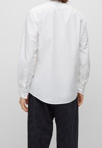 Vit långärmad skjorta med klassisk krage, med knäppningar i ärmsluten och rundad nederkant. Tillverkad av slätt tyg med subtil textur.