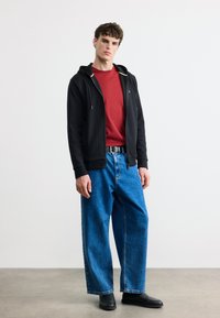 Νεαρός άνδρας που φοράει μαύρη ζακέτα με φερμουάρ, κόκκινη κοντομάνικη μπλούζα, φαρδιά μπλε τζιν, μαύρη ζώνη και μαύρα παπούτσια, στέκεται μπροστά σε λευκό φόντο.