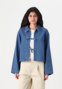 Izgriezta džinsa jaka zilā krāsā, ar pogām nesaturošu dizainu, kurā ir lentas, platām piedurknēm un tīru apdari. Valkāta virs baltas blūzes ar punktiņiem.