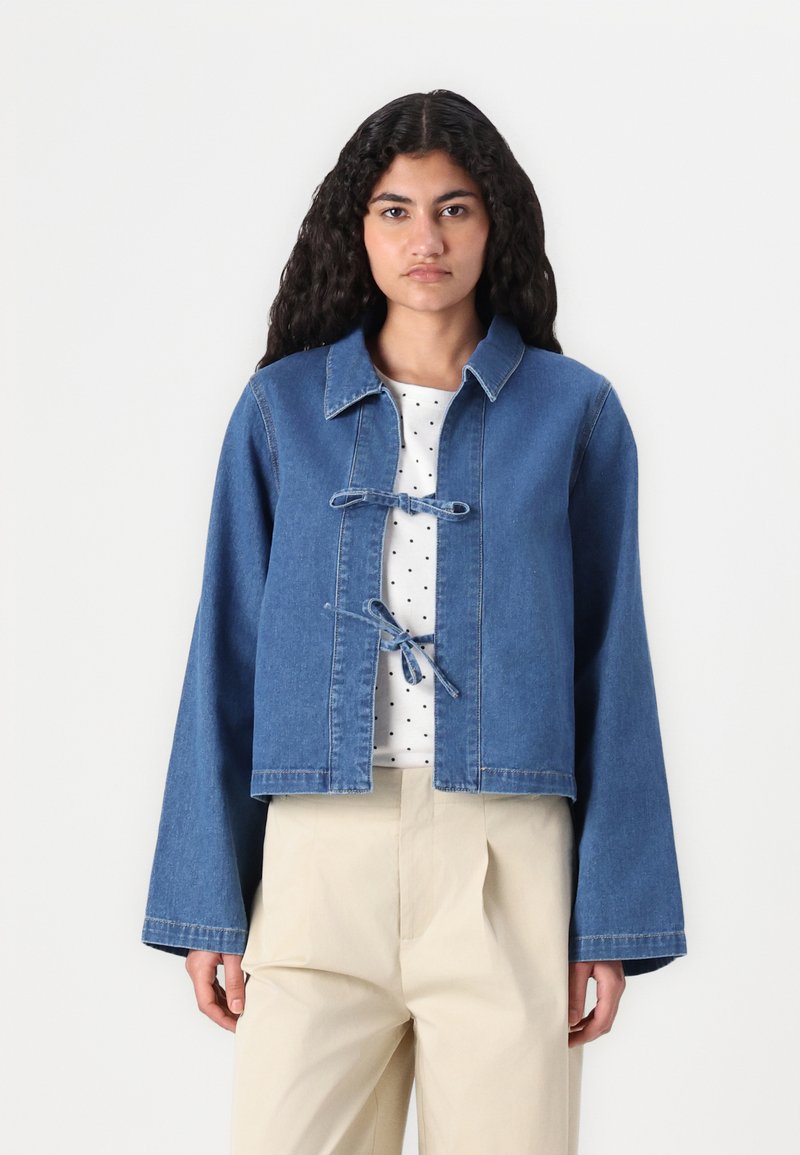 Izgriezta džinsa jaka zilā krāsā, ar pogām nesaturošu dizainu, kurā ir lentas, platām piedurknēm un tīru apdari. Valkāta virs baltas blūzes ar punktiņiem.