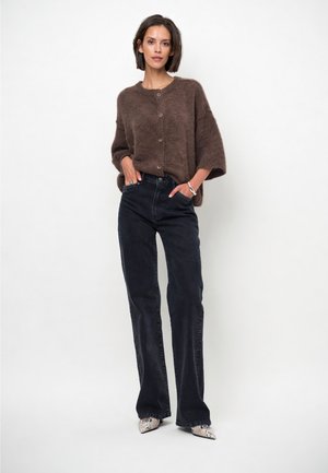 Vrouw met een bruine zachte gebreide vest met knopen, zwarte high-waist wijde pijpen jeans en puntige schoenen met slangenprint, staand met handen in de zakken.