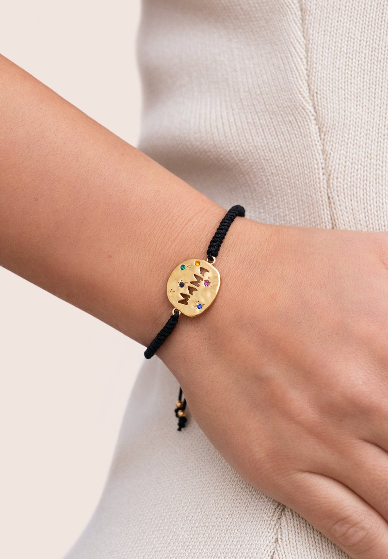 Pulsera de charm ovalado de oro sobre un cordón negro, que presenta piedras preciosas coloridas y un acabado texturizado. Diseño minimalista, ajustable con nudos.