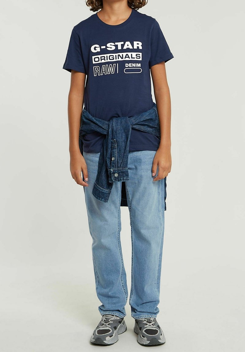 Enfant portant un t-shirt bleu marine G-Star Originals, un jean bleu clair, des baskets grises et une veste en jean nouée autour de la taille.