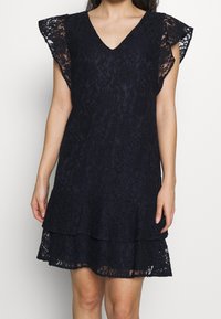 Marinblå spetsklänning med V-ringning, korta volangärmar och ett tiersat nederkant. Tyget har ett blommönster och en strukturerad yta.