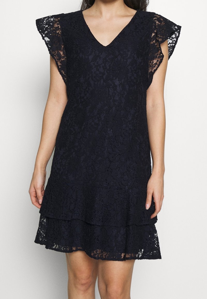 Marinblå spetsklänning med V-ringning, korta volangärmar och ett tiersat nederkant. Tyget har ett blommönster och en strukturerad yta.