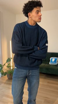 Μπλε ναυτικό ριγέ πλεκτό πουλόβερ με ριμπ υφή και ψηλό λαιμό, σε συνδυασμό με ανοιχτόχρωμο τζιν παντελόνι. Σημαντικές λεπτομέρειες περιλαμβάνουν μακριά μανίκια και ετικέτα έμφασης.