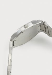 Montre en acier inoxydable avec un boîtier octogonal, texture brossée et bracelet à maillons. Présente une couronne proéminente et un fond gravé.