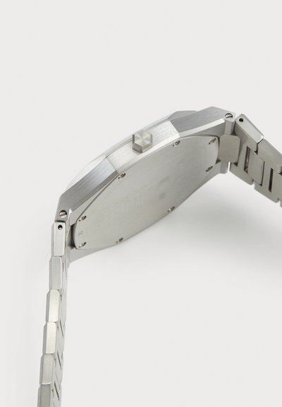 Rozsdamentes acél óra nyolcszögletű tokkal, kefezett felülettel és láncos karkötővel. Kiemelkedő koronával és gravírozott hátlappal rendelkezik.