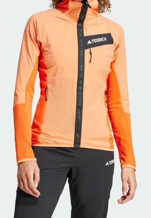 Person trägt eine orange Terrex-Jacke mit schwarzem Reißverschluss und schwarze Terrex-Hose, steht mit entspannten Händen an den Seiten.