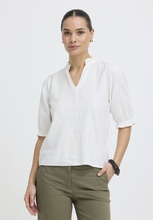 Femme aux cheveux foncés portant un chemisier blanc à col en V et manches volantées, un pantalon vert olive, des boucles d'oreilles dorées et un bracelet noir.