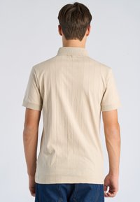 Mann trägt beige kurzärmeliges Poloshirt mit dünnen vertikalen Streifen, von hinten zu sehen, steht vor einem einfachen hellen Hintergrund.
