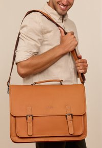 Tan leather messenger bag with a structured shape. Features a top handle, adjustable strap, and buckle accents. Smooth texture.