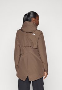 Een bruine waterdichte jas met een capuchon, uitgerust met een rugflap, elastische manchetten en een logo aan de bovenlinkerkant. Gladde textuur en een middenlange stijl.