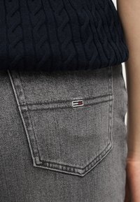 Close-up of gray denim jeans pocket with a small rectangular logo and a navy blue cable knit sweater above.