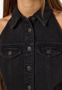 Černé džínové šaty bez rukávů s knoflíkovým zapínáním vpředu, špičatým límcem a dvěma náprsními kapsami. Obsahují kovové knoflíkové detaily a texturovanou tkaninu.