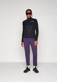 Fekete cipzáras dzsekit, lila nadrágot, fekete sportcipőt és túlméretezett fekete napszemüveget viselő nő áll egy sima fehér háttér előtt.