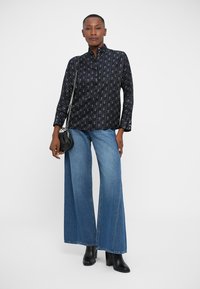Zwarte blouse met knoopsluiting en een geometrisch patroon, gecombineerd met brede blauwe spijkerbroek. Model houdt een kleine zwarte handtas vast.