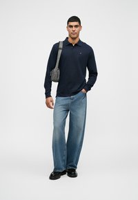 Polo de manga comprida azul-marinho com um pequeno logótipo, combinado com jeans de ganga azul-claro de perna larga. Sapatos pretos e uma mala de ombro cinzenta.