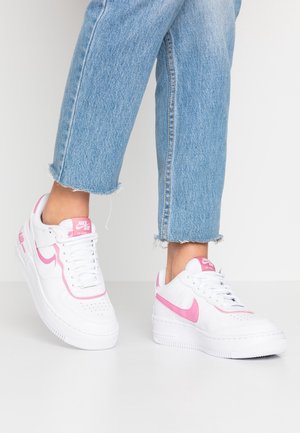 Osoba ubrana w jasnoniebieskie krótkie dżinsy i białe buty Nike Air Force 1 z różowym logo i detalami na białej podłodze.
