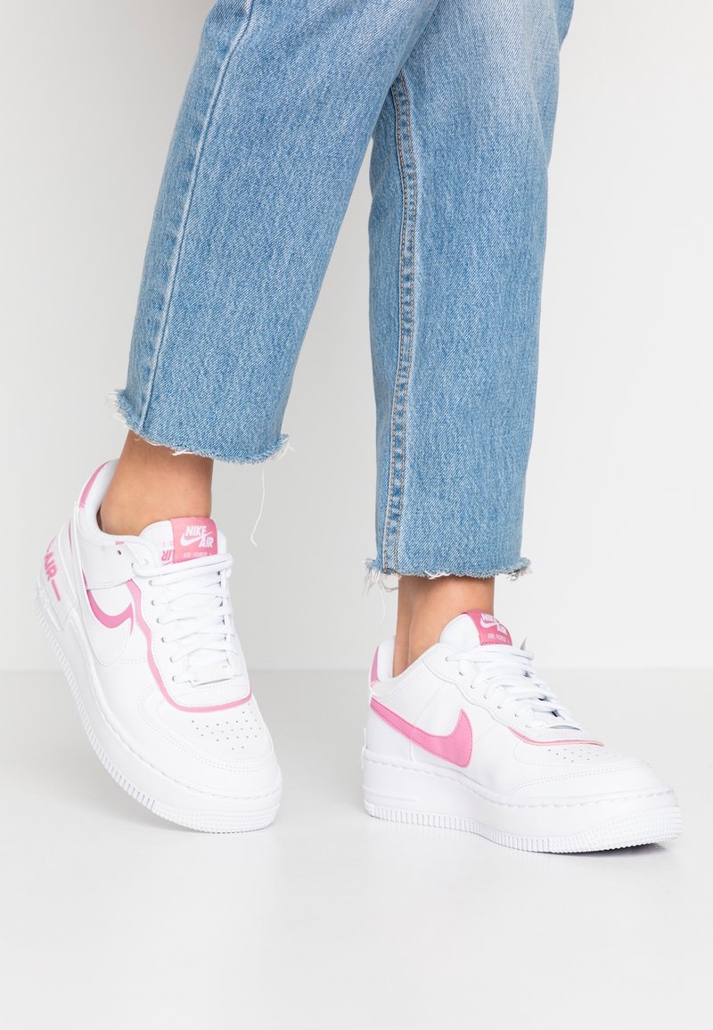 Personne portant un jean court bleu clair et des baskets blanches Nike Air Force 1 avec un swoosh rose et des détails sur un sol blanc.