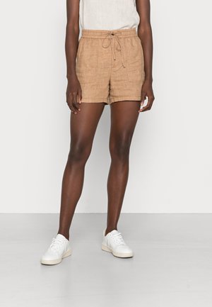 Personne portant un short beige clair à cordon, un haut blanc texturé et des baskets blanches, debout devant un fond uni clair.