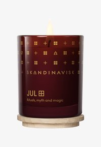 Bougie rouge allumée dans un pot en verre avec des symboles nordiques dorés, le texte "JUL" et "Skandinavisk", placée sur un sous-verre en bois rond.