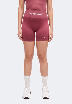 Person iført mørkerøde Nike Pro tætsiddende shorts, hvide crew-sokker og hvide Nike-sneakers stående mod en ensfarvet baggrund.