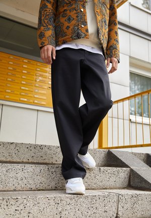 Schwarze weit geschnittene Hosen und ein gemustertes braunes Hemd werden mit einem weißen Schichtoberteil und weißen Turnschuhen kombiniert, während sie auf strukturierten Betontreppen stehen.