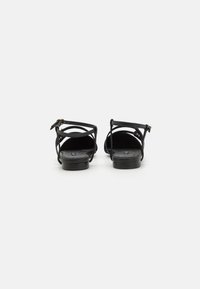 Black strappy sandals with a flat sole, featuring a buckle strap and crisscross design on the upper. Smooth texture and minimalist style.
