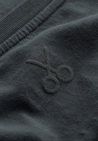 Donkergrijze stof met een gestructureerde oppervlakte heeft een verhoogd, tonale schaarlogo. De stof heeft een gladde, zachte uitstraling.