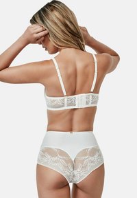 Femme vue de dos ajustant ses cheveux, portant de la lingerie en dentelle blanche avec des motifs floraux sur le soutien-gorge et des culottes taille haute.