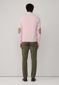 Uomo in piedi, di spalle, che indossa un maglione rosa chiaro con toppe marroni sui gomiti, pantaloni cargo oliva e scarpe marroni scure.