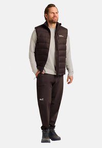 Zwarte gewatteerde bodywarmer over een lichtgrijs shirt met lange mouwen, gecombineerd met donkere broek en zwarte wandel- of bergschoenen, staand voor een neutrale achtergrond.