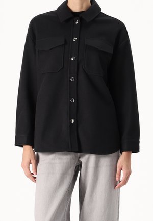 Chaqueta negra abotonada con bolsillos en el pecho, usada sobre unos vaqueros gris claro, mostrada desde los hombros hasta las caderas sobre un fondo blanco.